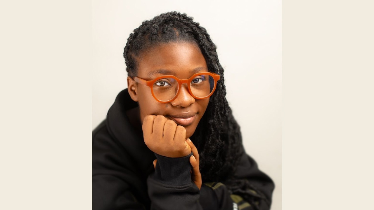 A picture showing Motun in a hooded jacket. Motun wedges her chin on her fist, staring at the camera with oversized glasses.