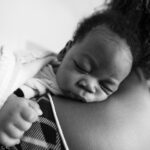 A monochrome image of a male infant sleeping soundly on his mothers shoulders.