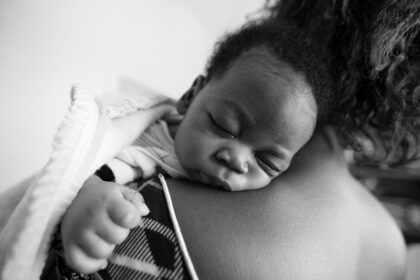 A monochrome image of a male infant sleeping soundly on his mothers shoulders.
