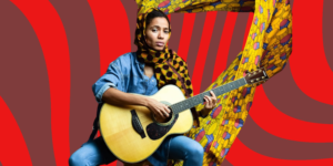 A woman in demin straddles a guitar while staring intently at the camera. Her head is turbanned in Ankara, and her background is coloured by a flowing Ankara material.