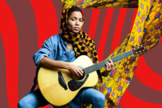 A woman in demin straddles a guitar while staring intently at the camera. Her head is turbanned in Ankara, and her background is coloured by a flowing Ankara material.